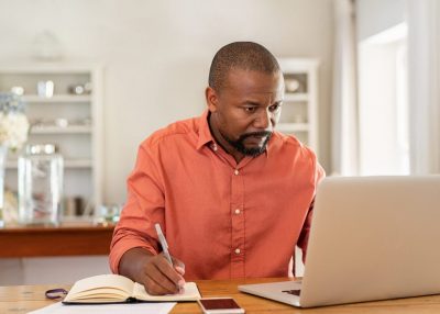 Mature man working on laptop at home Mature man working on laptop while taking notes. Businessman working at home with computer while writing on agenda. African man managing home finance, reviewing bank account and using laptop in living room.