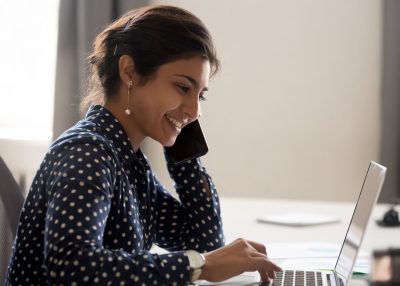 Smiling employee working on laptop talking over phone Smiling female employee multitask working on computer in office talking over phone, happy woman worker using laptop, laughing speaking with friend on cell or communicating with client online